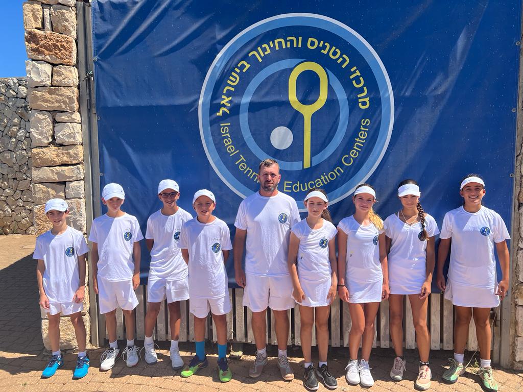 Four boys, their coach, and four girls stand in front of ITEC logo on blue background. Purpose of image is to show the kids from Israel praticipating in tennis exchange. 