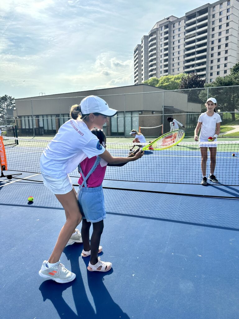 12 year old girl helps young girl learn how to hold tennis racket during afterschool clinic